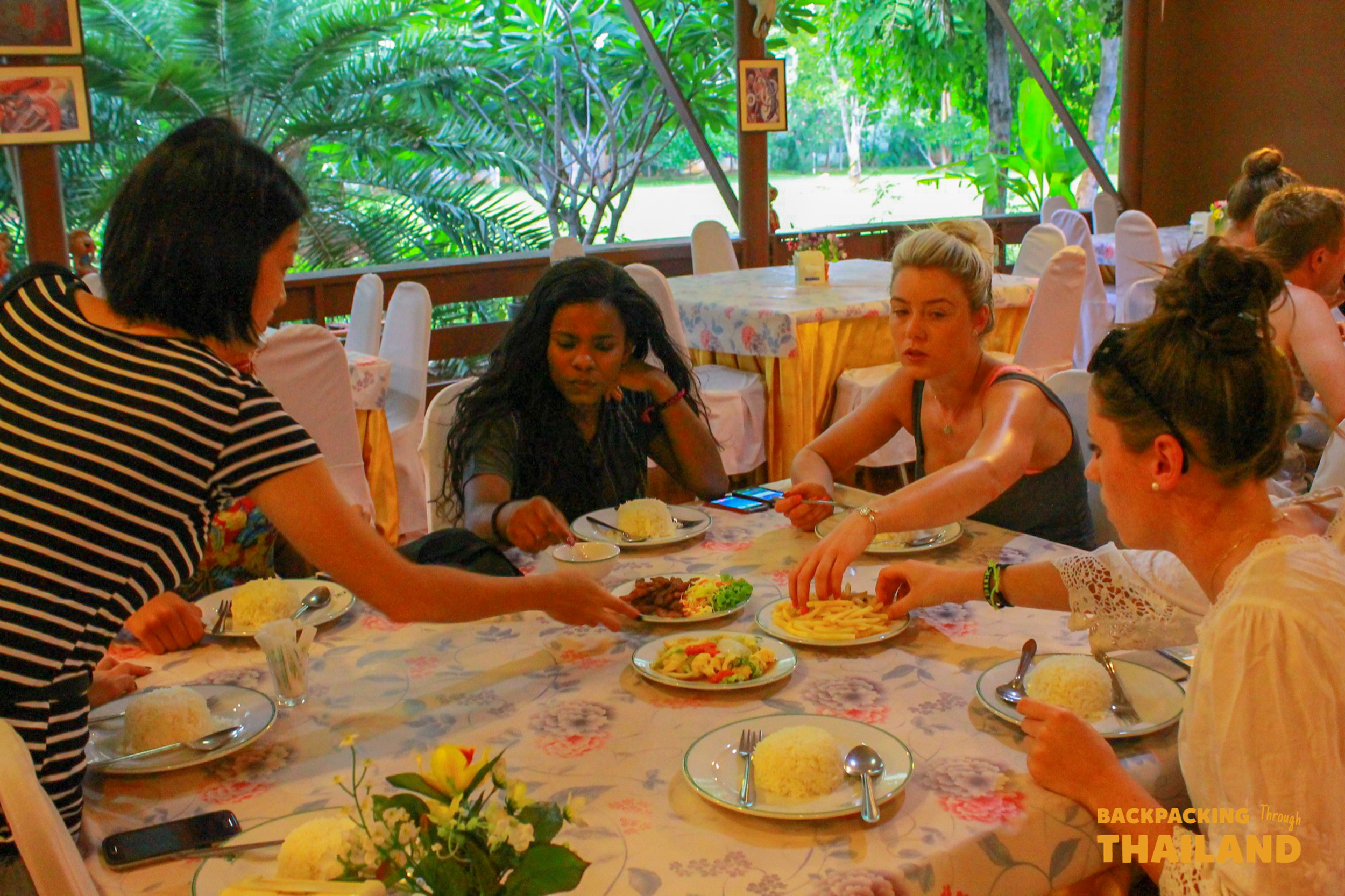 Backpackers enjoying a group meal with traditional dishes at a resort in Singburi, Thailand.