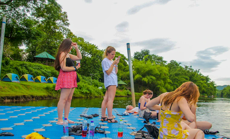 Backpackers relax on a floating platform by the Khwae Yai River with colorful tents on the bank.