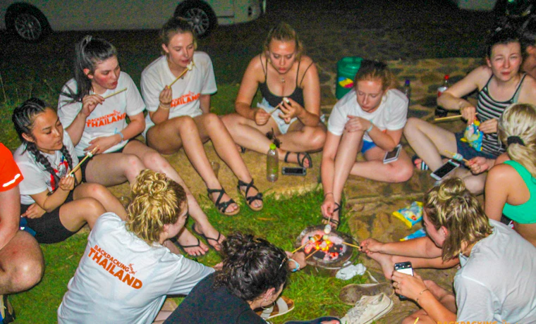 Backpackers gather around a campfire, roasting marshmallows and enjoying a social evening at Erawan National Park.