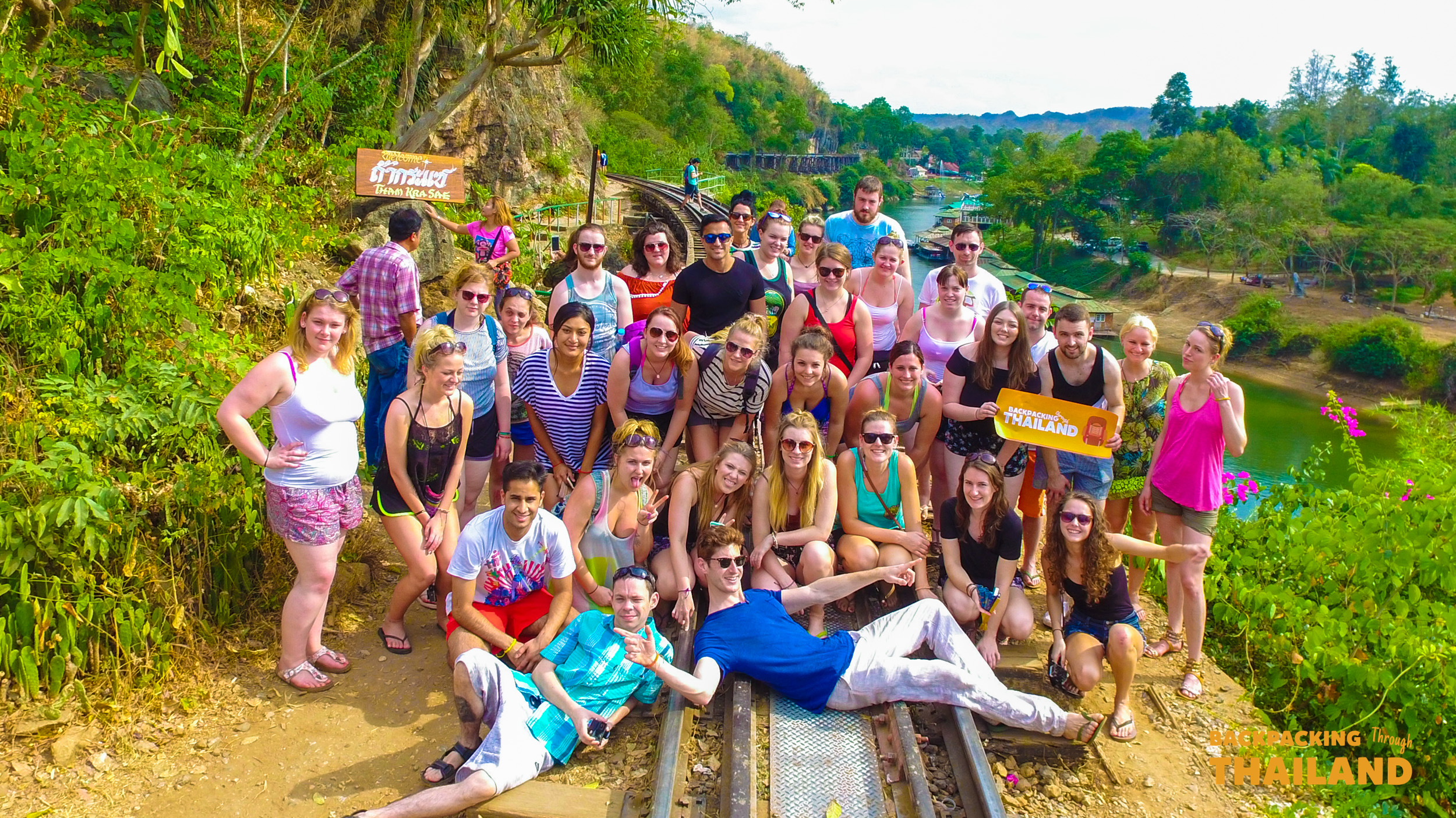 A large group of happy backpackers pose on old railway tracks in Erawan National Park, Thailand.