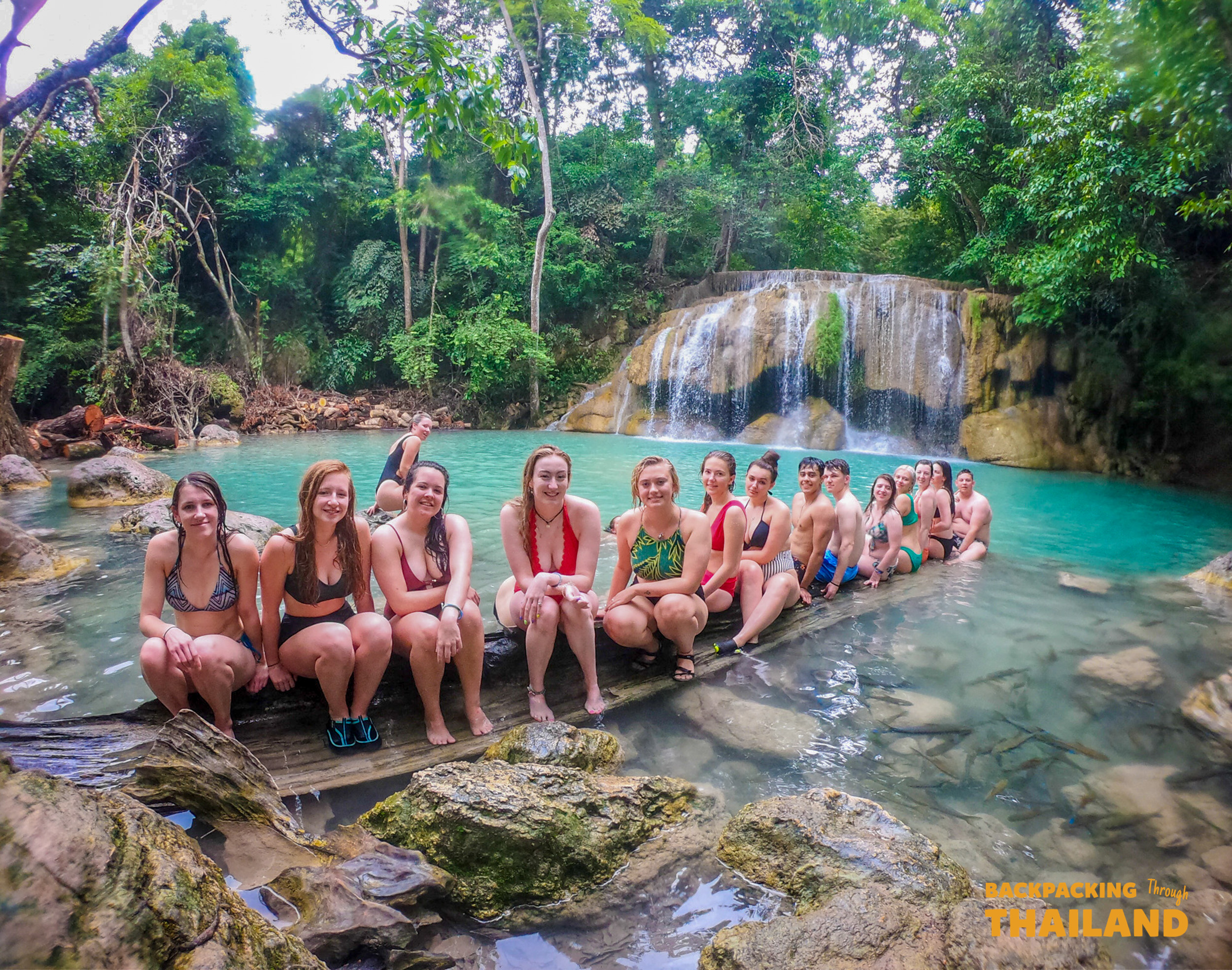 Backpacking group sitting on a wooden platform beside a turquoise waterfall pool at Erawan National Park