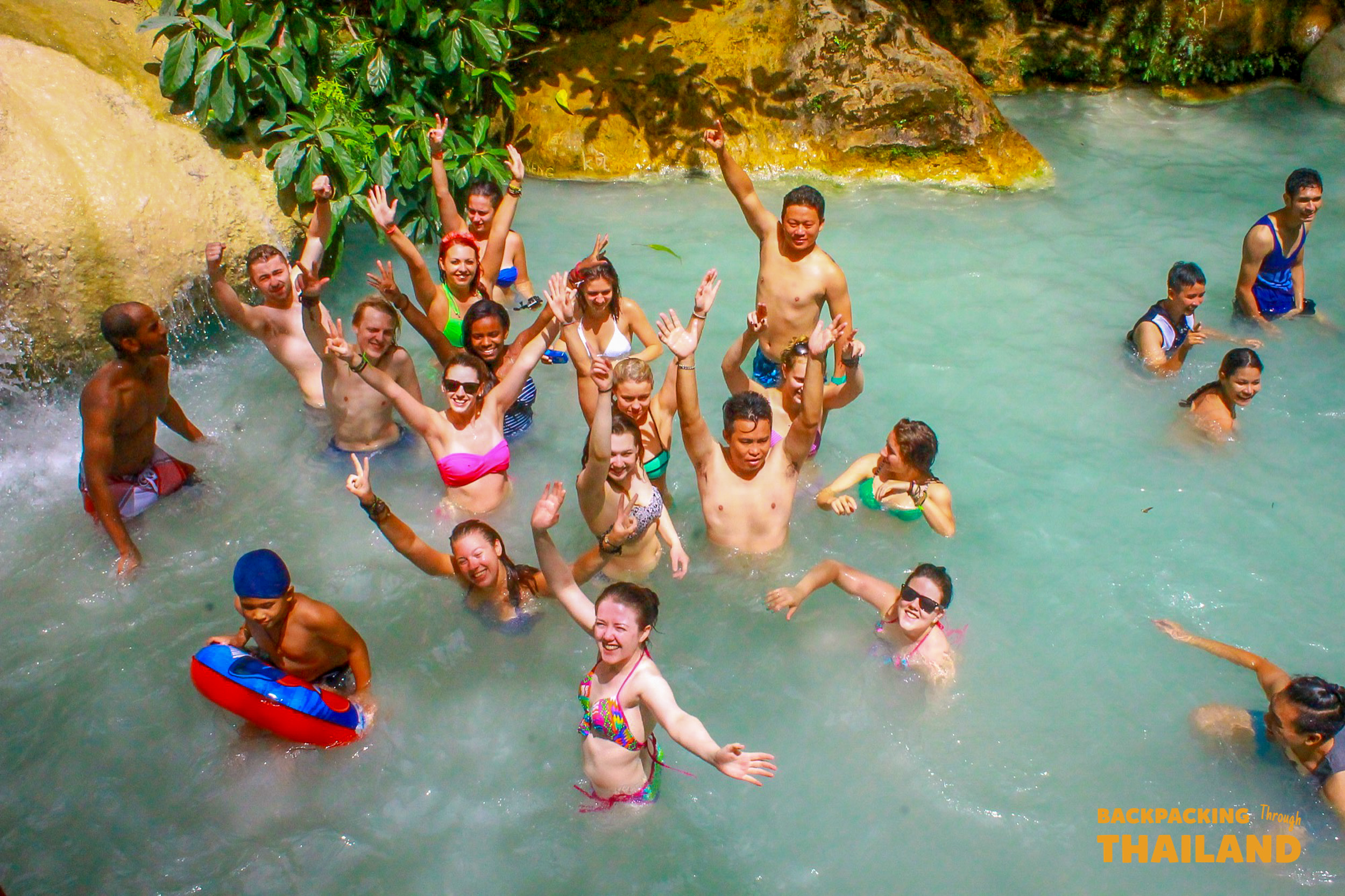 Joyful backpackers raise their arms while swimming in the refreshing emerald waters of Erawan National Park.