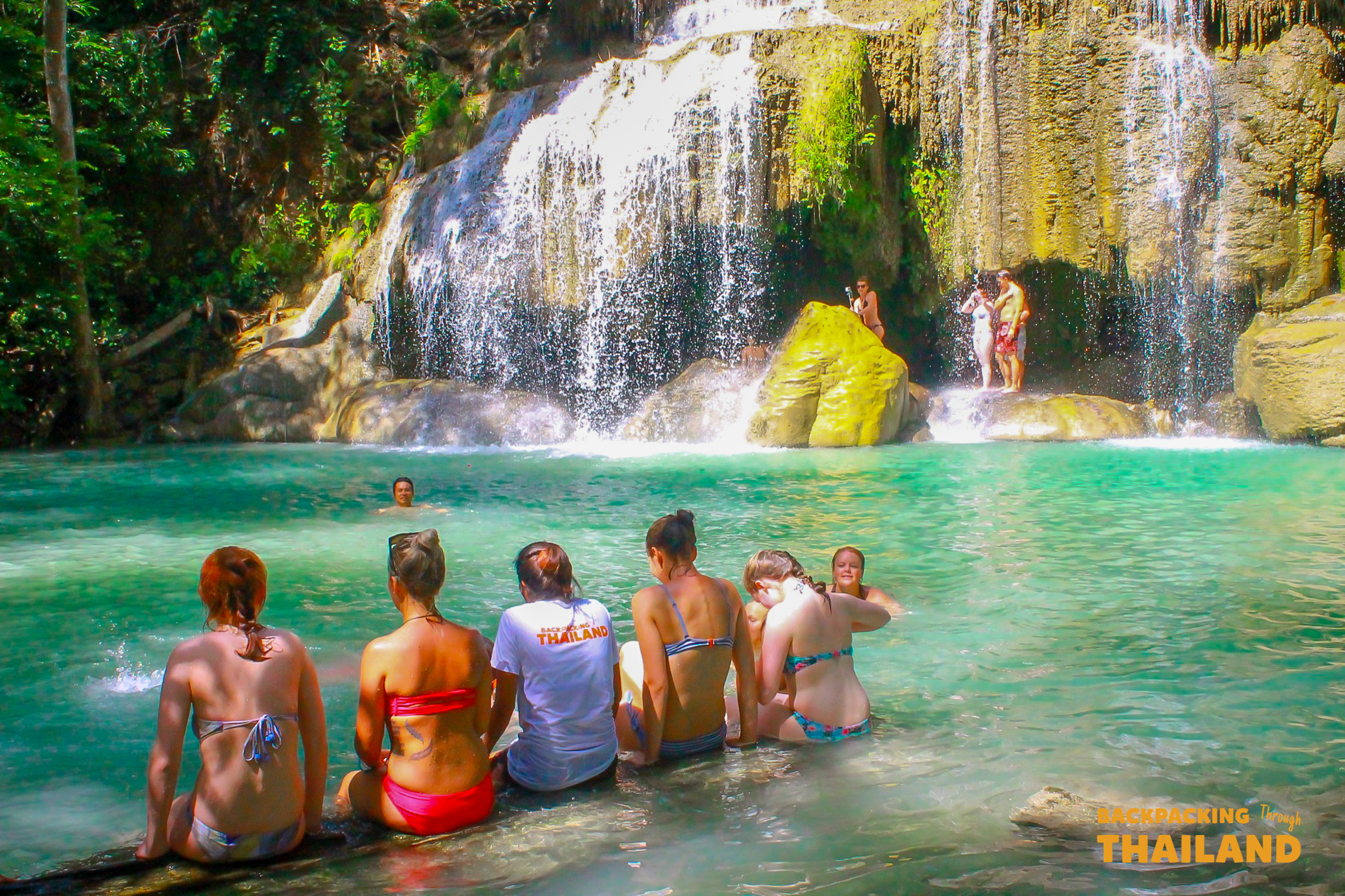 Backpacking group relaxing in a turquoise waterfall pool with a cascading waterfall in the background at Erawan National Park