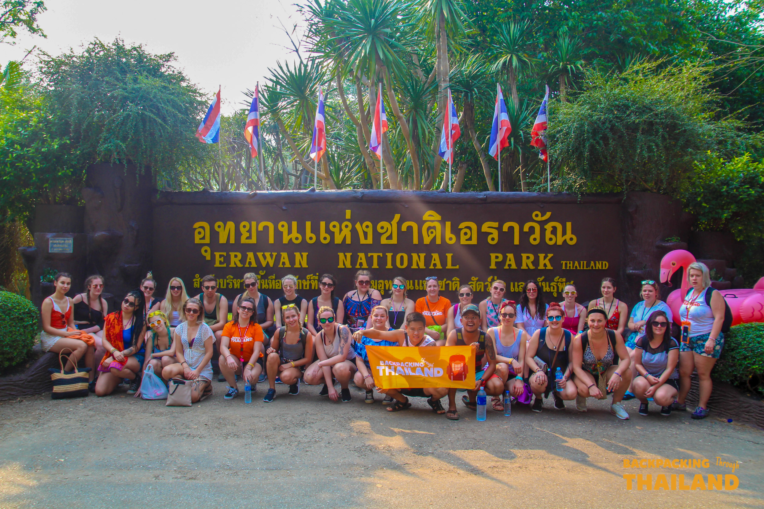 Backpacking group posing in front of the Erawan National Park sign with flagsBackpacking group relaxing in a turquoise waterfall pool with a cascading waterfall in the background at Erawan National Park
