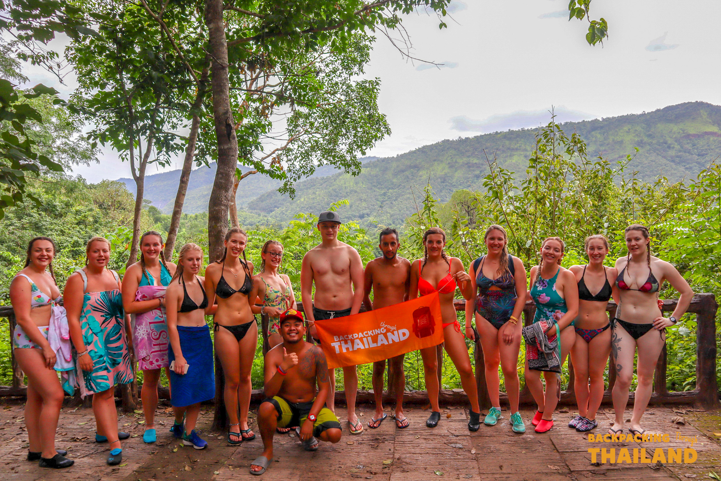 Backpacking group posing together on a bench at Erawan National Park with green hills in the background