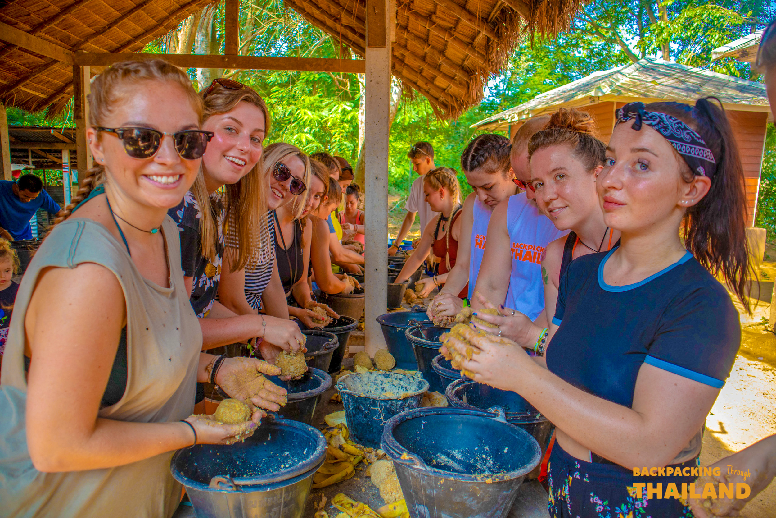 Smiling backpackers prepare food for elephants in buckets at a sunny Thai nature park.