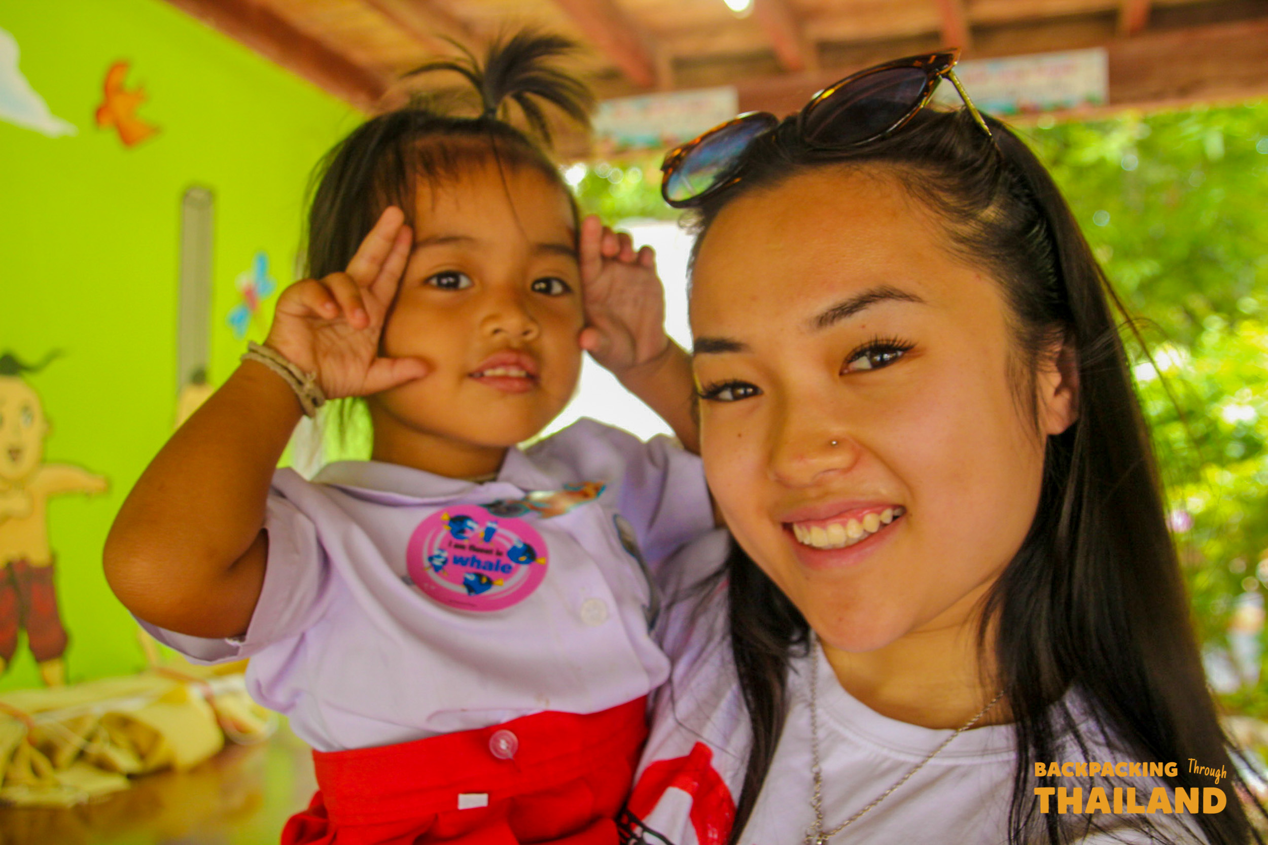 Backpacking traveller holding a Thai child and smiling at the camera at the schoolBackpacking group and local schoolchildren sitting in a colorful classroom, chatting and smiling