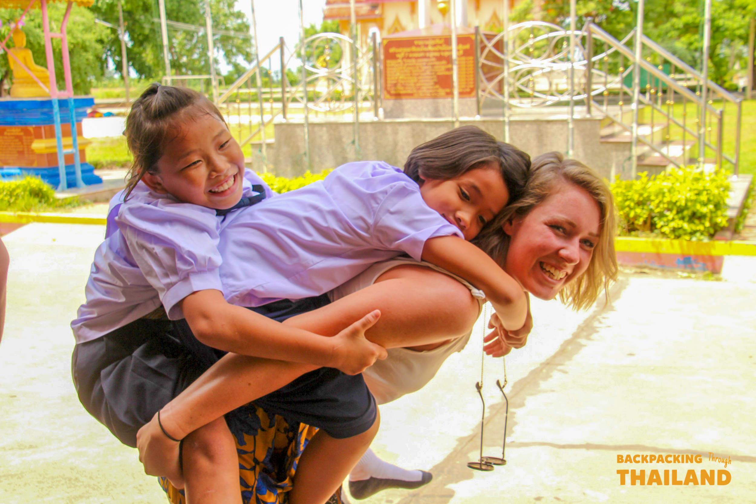 Backpacking travellers playing with Thai children, giving piggyback rides and laughing together at the school