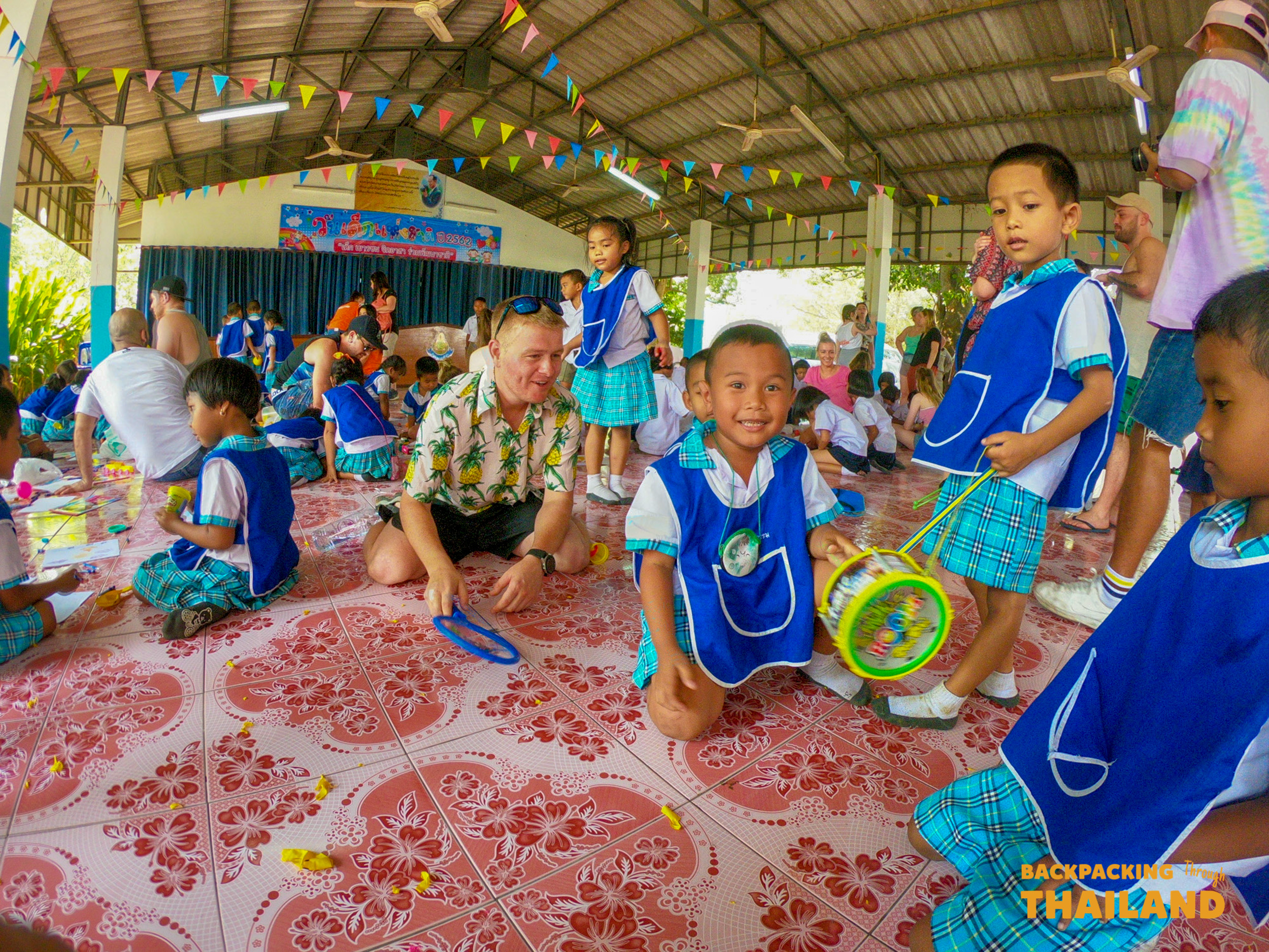 A backpacker plays with smiling Thai school children on a tiled floor during a heartwarming visit in Thailand.