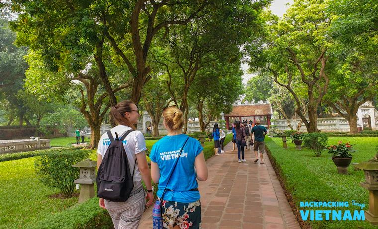 Two backpackers stroll through a beautiful, tree-lined garden with traditional architecture in Hanoi.