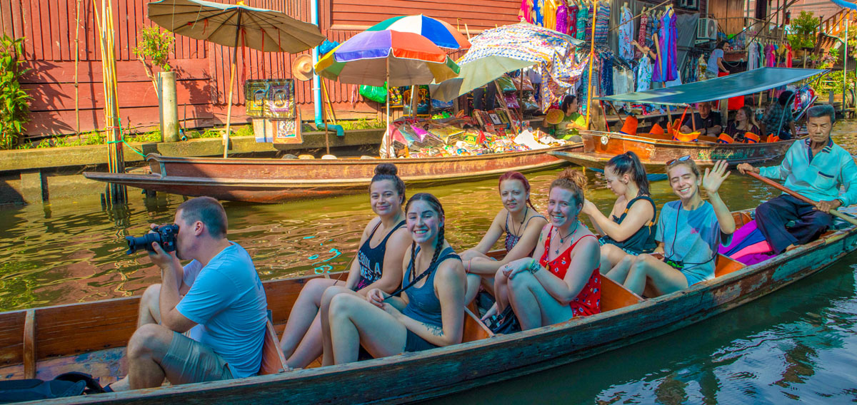 Smiling tourists on a traditional boat exploring a bustling floating market in Thailand with a local guide.