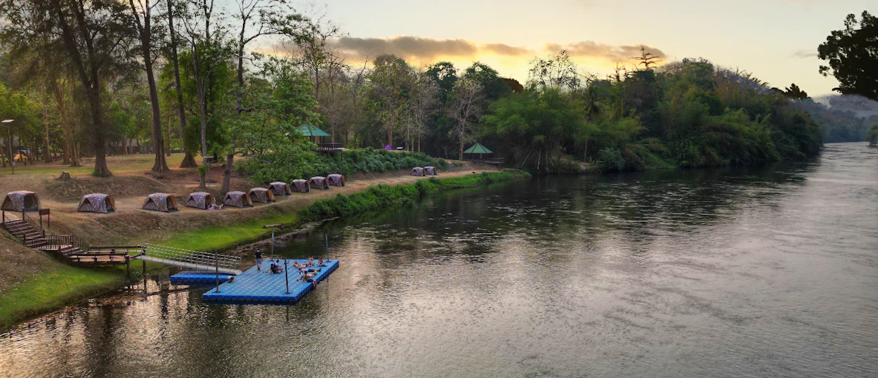 Tents pitched along the Khwae Yai River with a blue floating dock at sunset in Kanchanaburi, Thailand.