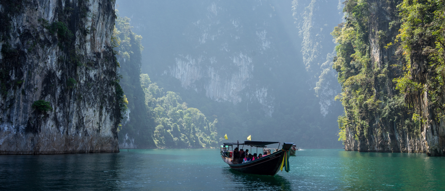 Long-tail boat floating on emerald Cheow Lan Lake between towering limestone cliffs and lush greenery