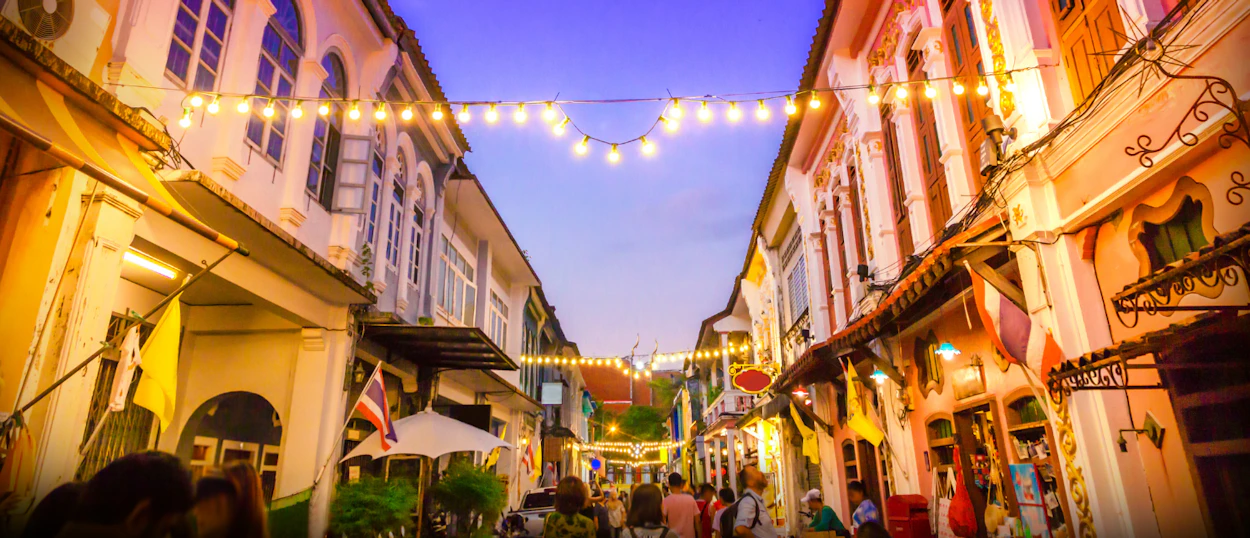 Colorful Sino-Portuguese buildings and string lights illuminate a lively street in Phuket Old Town at dusk.