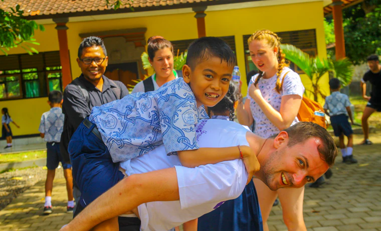 A joyful man gives a piggyback ride to a smiling boy in a sunny Bali village.