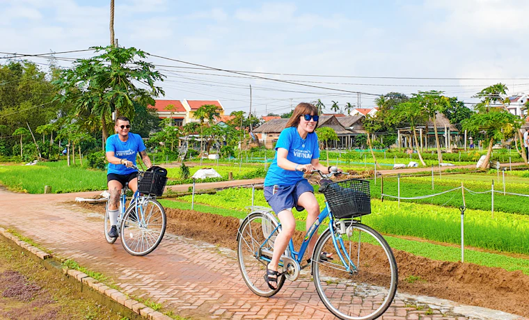 Two tourists happily cycle through lush green vegetable fields and traditional houses in Vietnam.