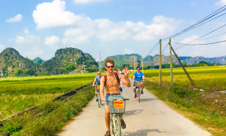 Three friends cycle on a sunny day through green rice paddies with dramatic limestone mountains in Vietnam.