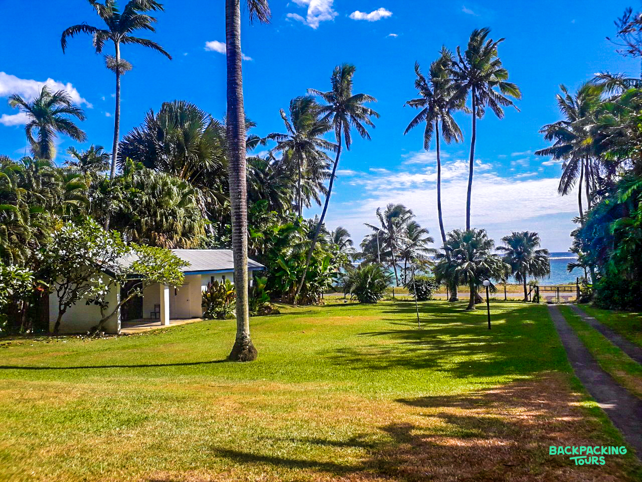 Lush tropical garden with a white building, tall palm trees, and a view of the sea.