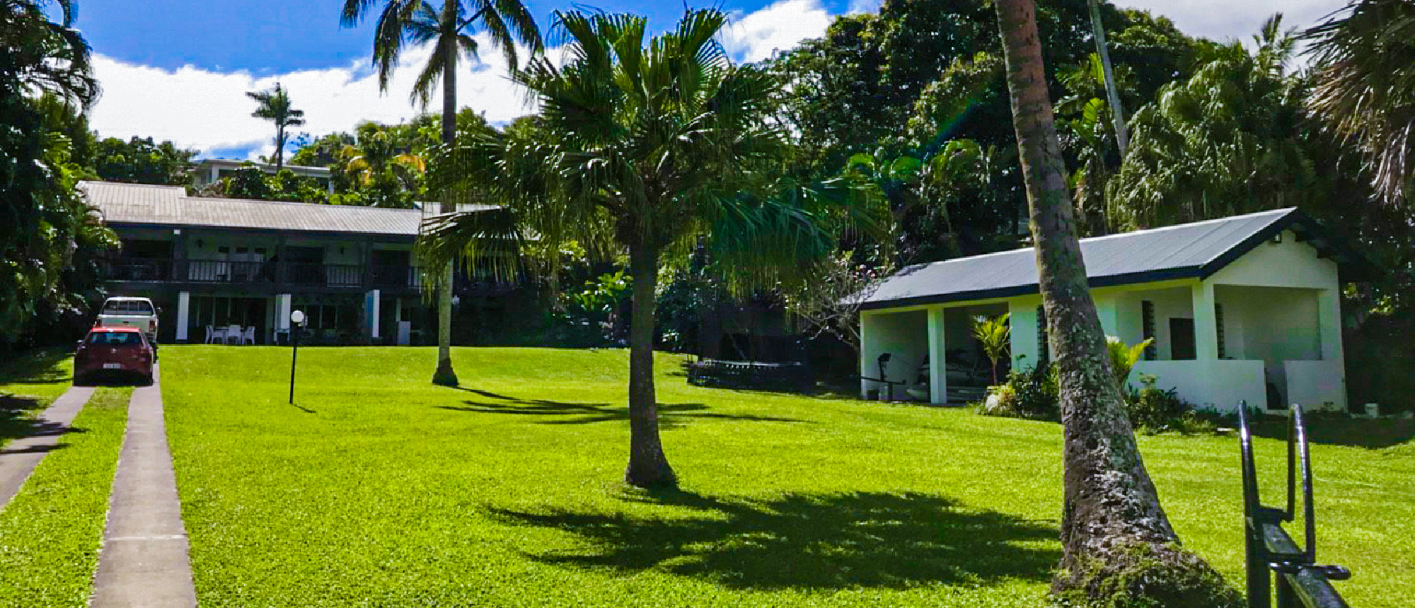 A peaceful tropical scene with a large green lawn, palm trees, and two buildings in Fiji.
