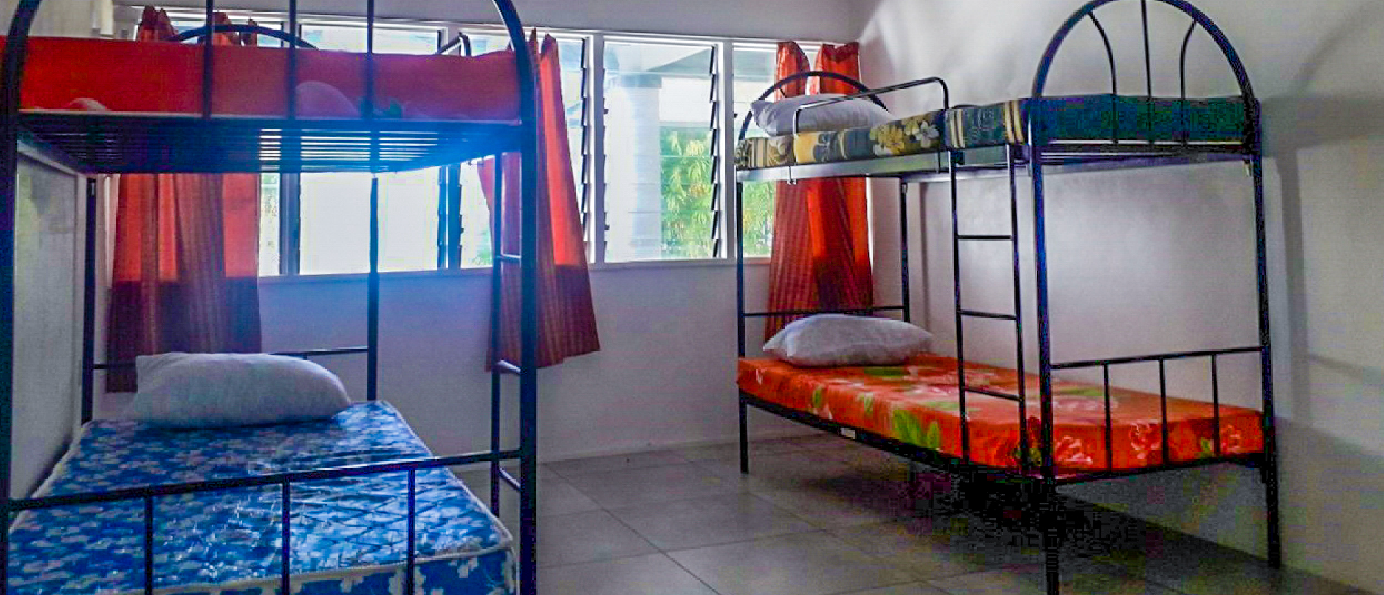 A simple dormitory room with two metal bunk beds, colorful mattresses, and red curtains.