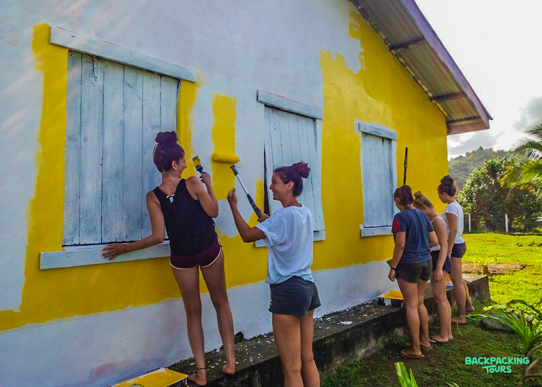 Young women volunteers painting a house yellow with blue shutters, surrounded by green foliage.