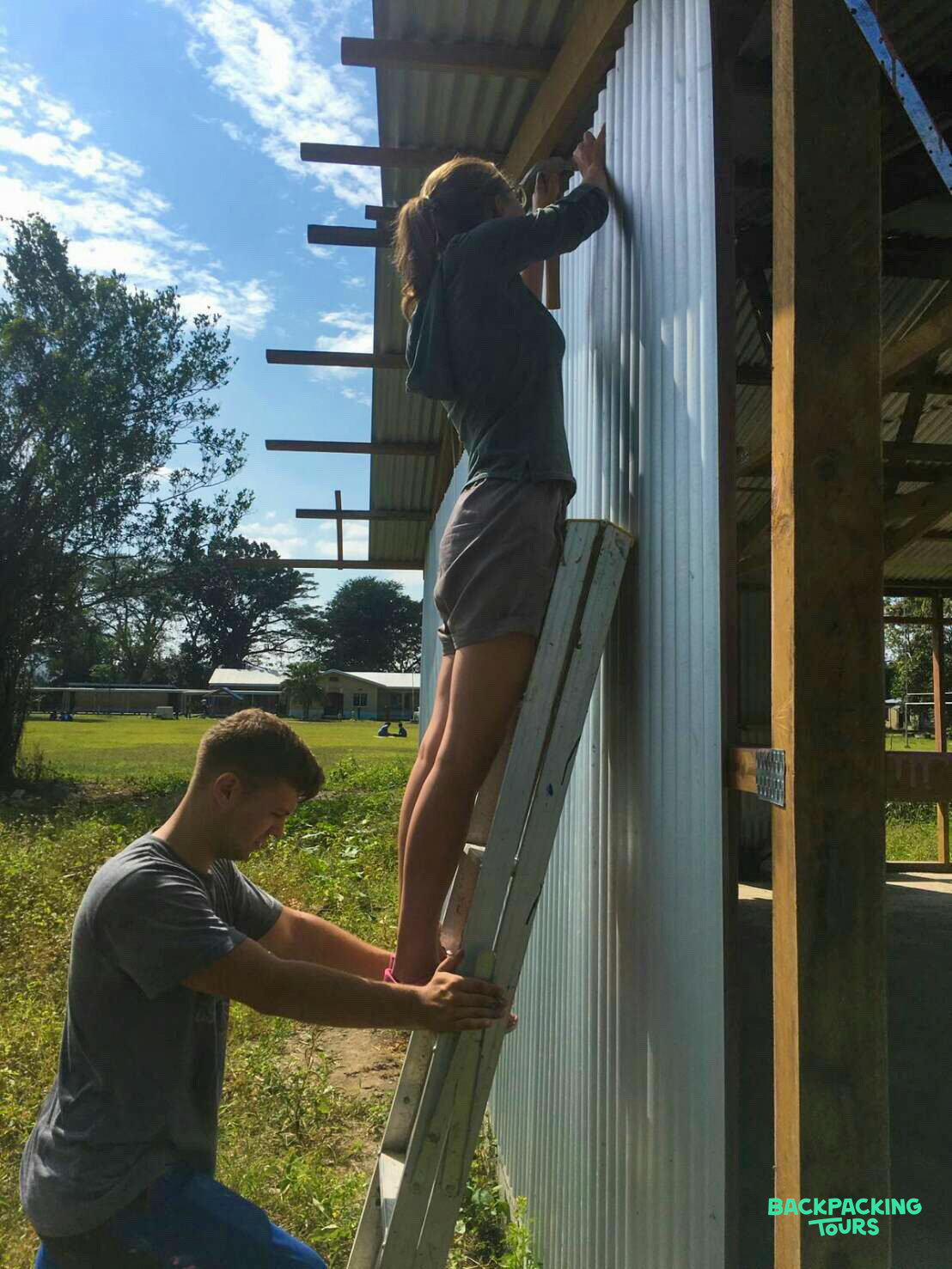 Two volunteers construct a building, one holding the ladder as the other installs metal siding.