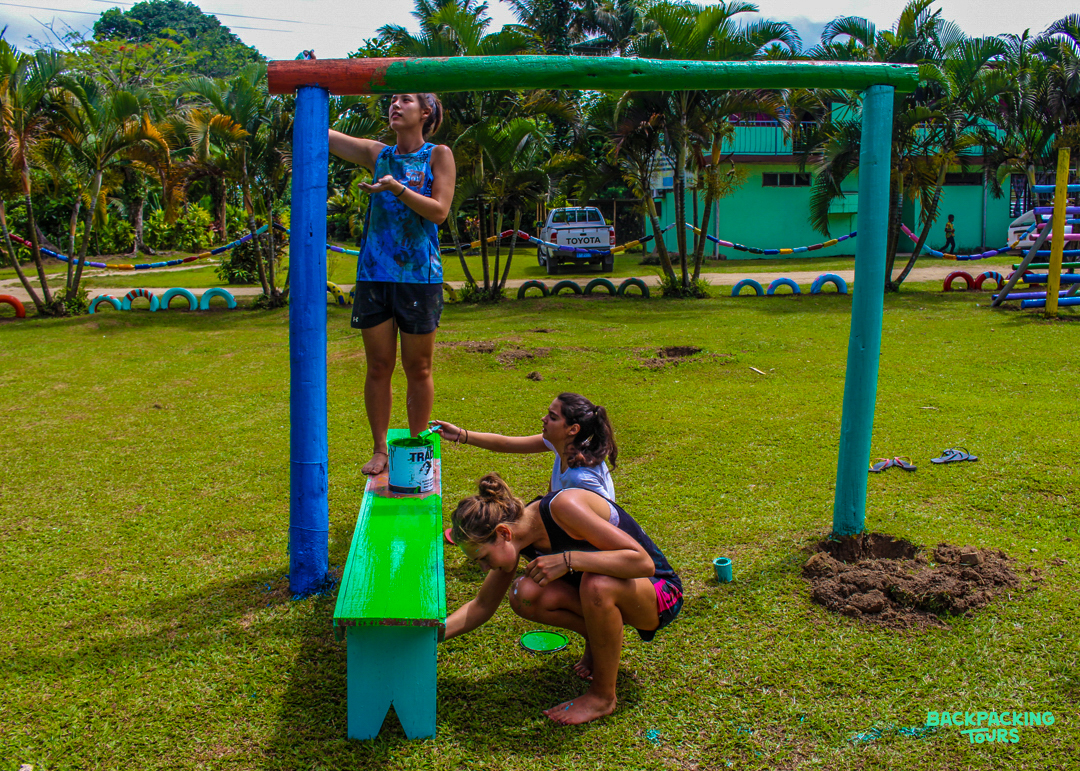 Three volunteers paint a colorful wooden playground structure on a sunny day in Fiji.