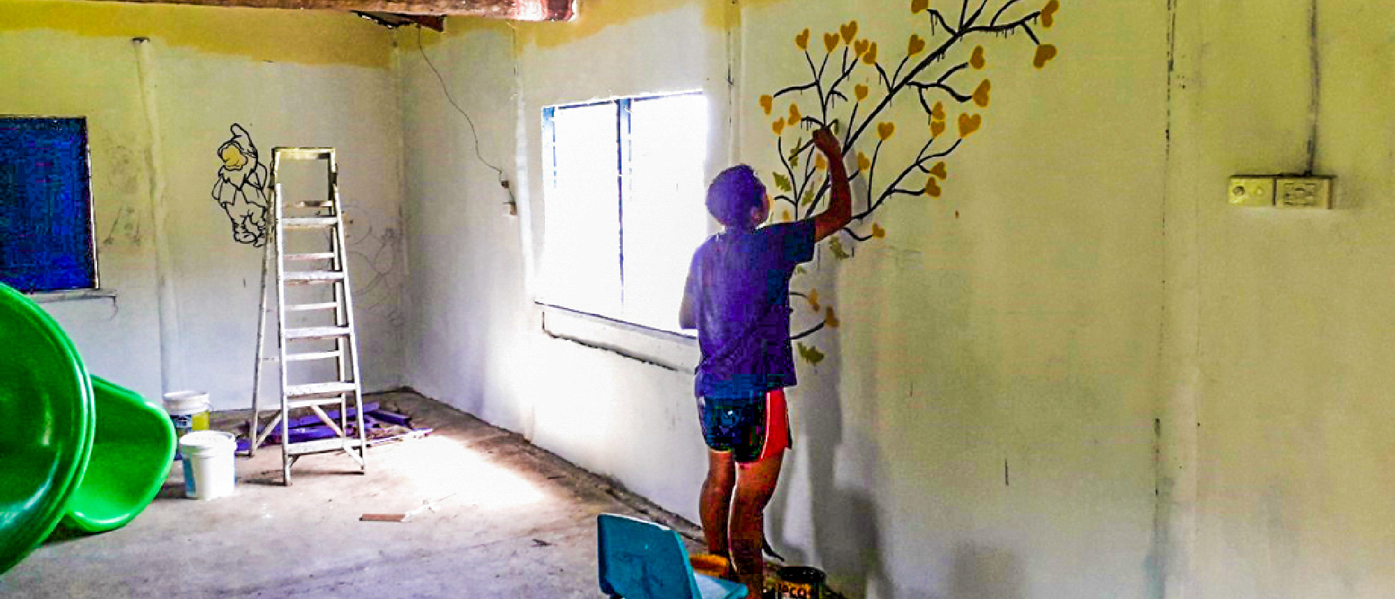 A person paints a tree mural with yellow heart-shaped leaves on a white wall.