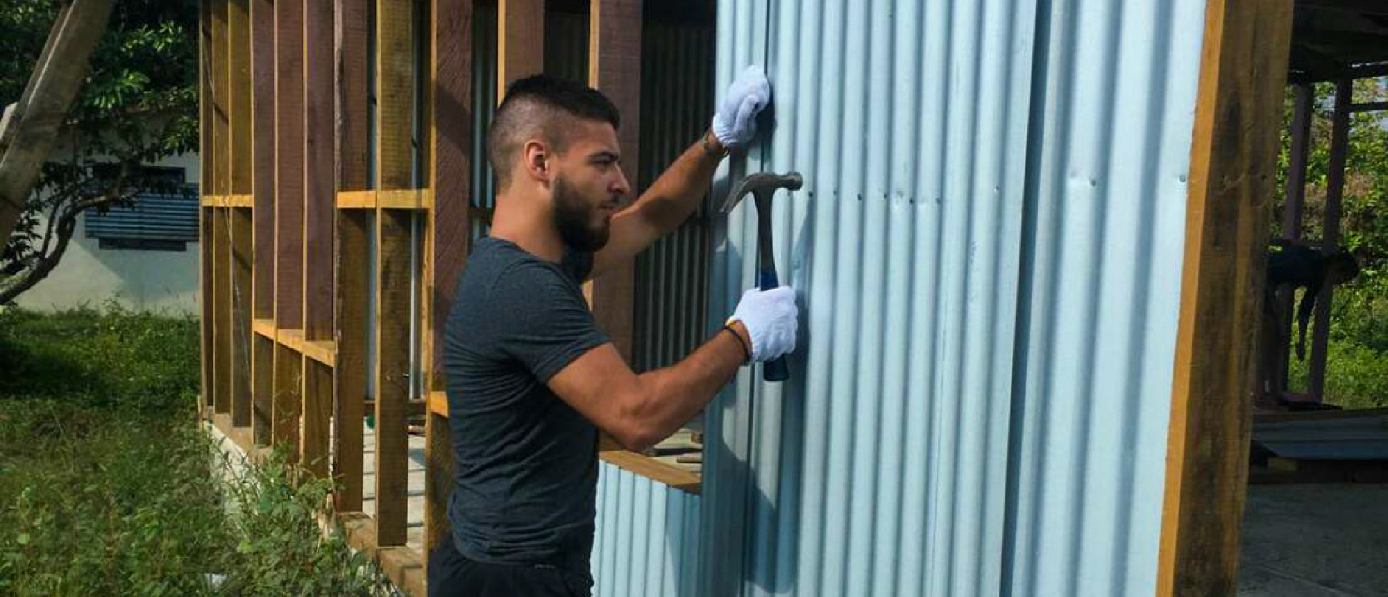 A man in white gloves hammers a light blue corrugated metal sheet onto a building frame.