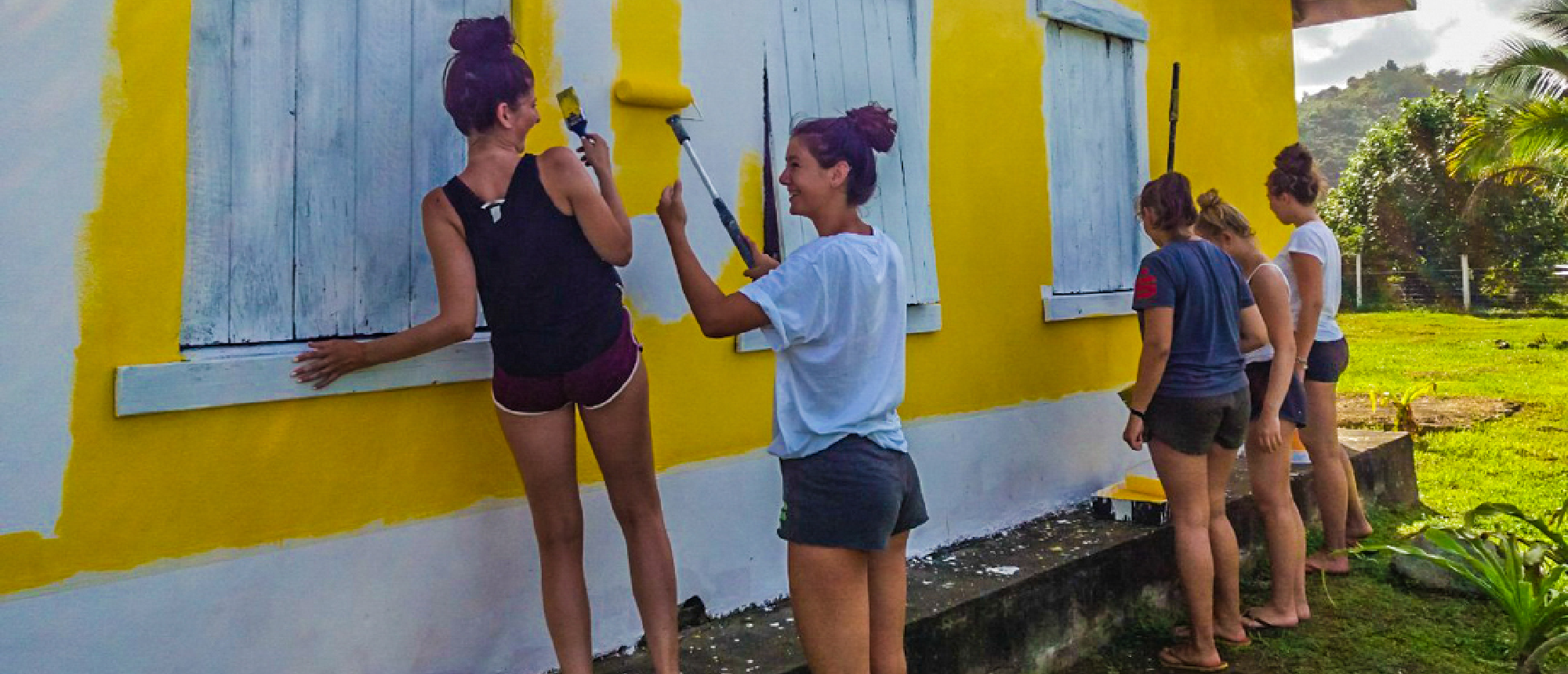 Smiling volunteers paint a vibrant yellow house wall with blue shutters in a lush outdoor setting.