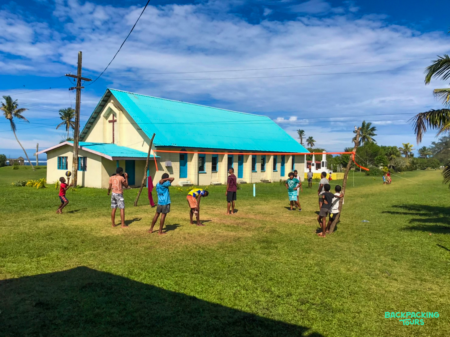 Children play a lively ball game on a sunny field outside a blue-roofed church in Fiji.