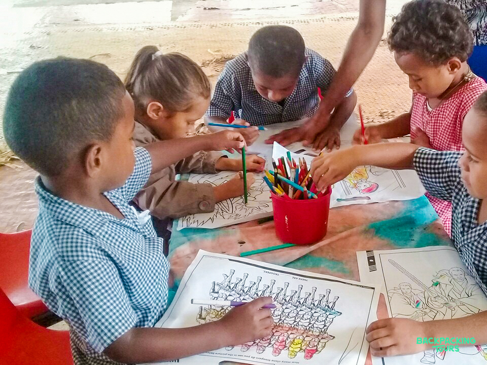 Young children in Fiji happily color pictures at a kindergarten table with an adult.