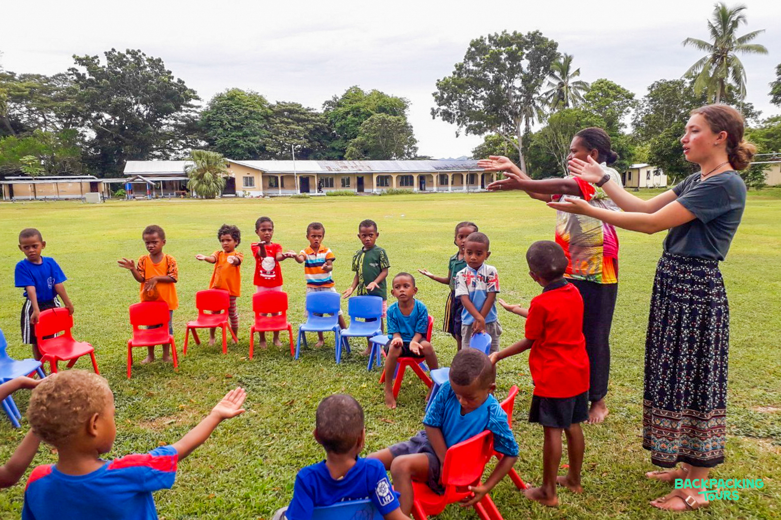 Volunteers lead a fun outdoor game with children in a circle of colorful chairs in Fiji.