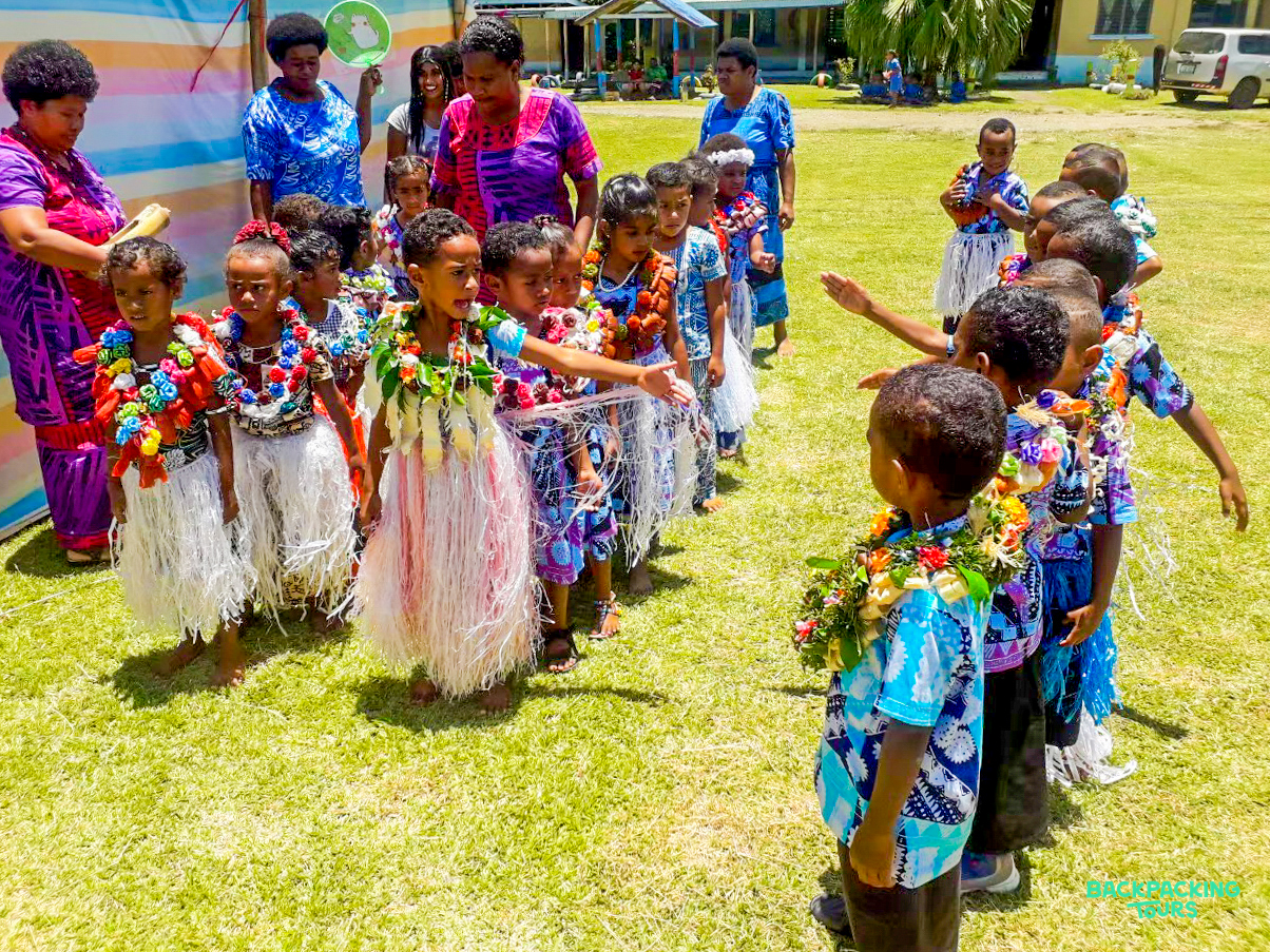 Fijian children in festive traditional attire and flower garlands participate in an outdoor community celebration.