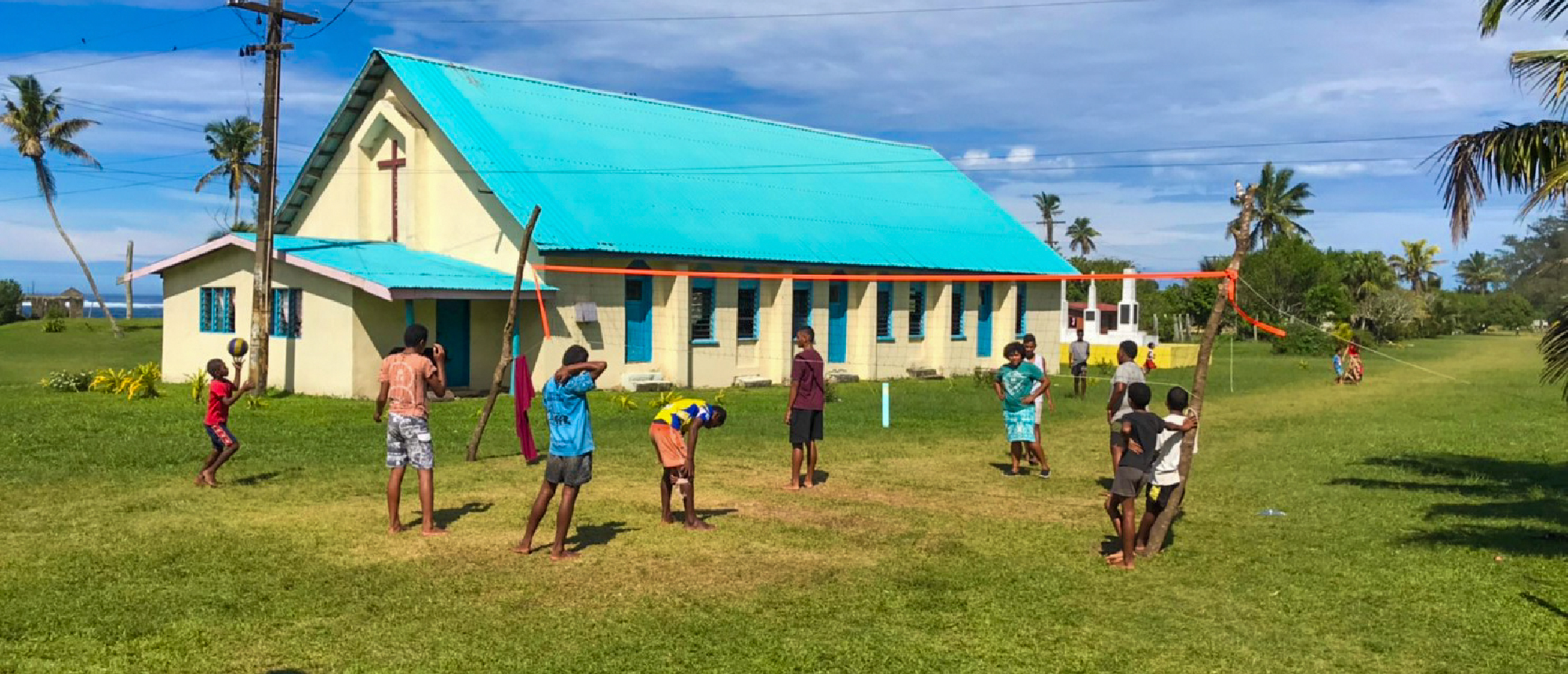 Children play a ball game on a sunny grassy field near a blue-roofed building in Fiji.