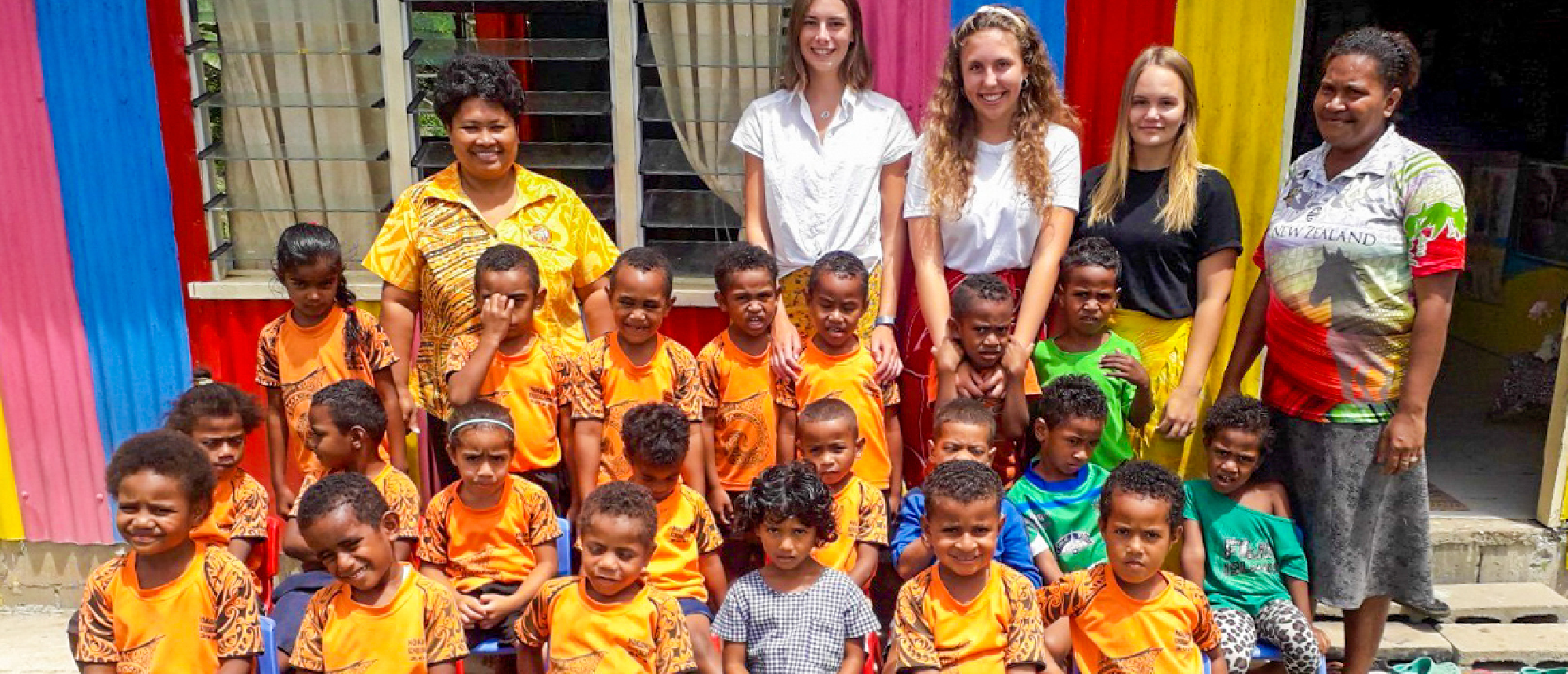 Volunteers and local kindergarten children smiling together in front of a vibrant building.