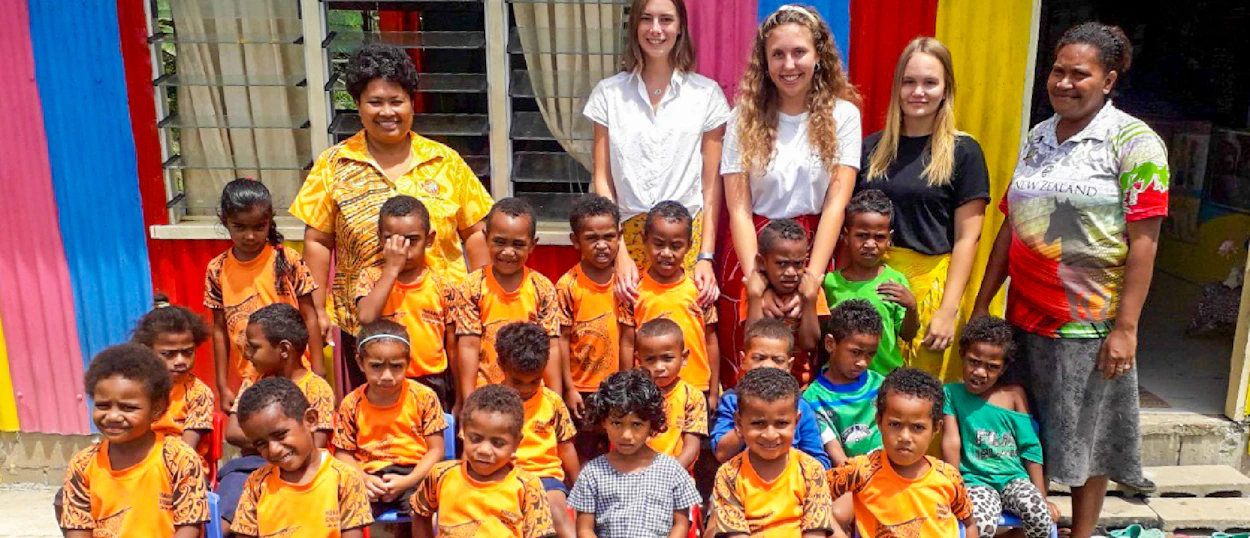 Volunteers and local kindergarten children smiling together in front of a vibrant building.