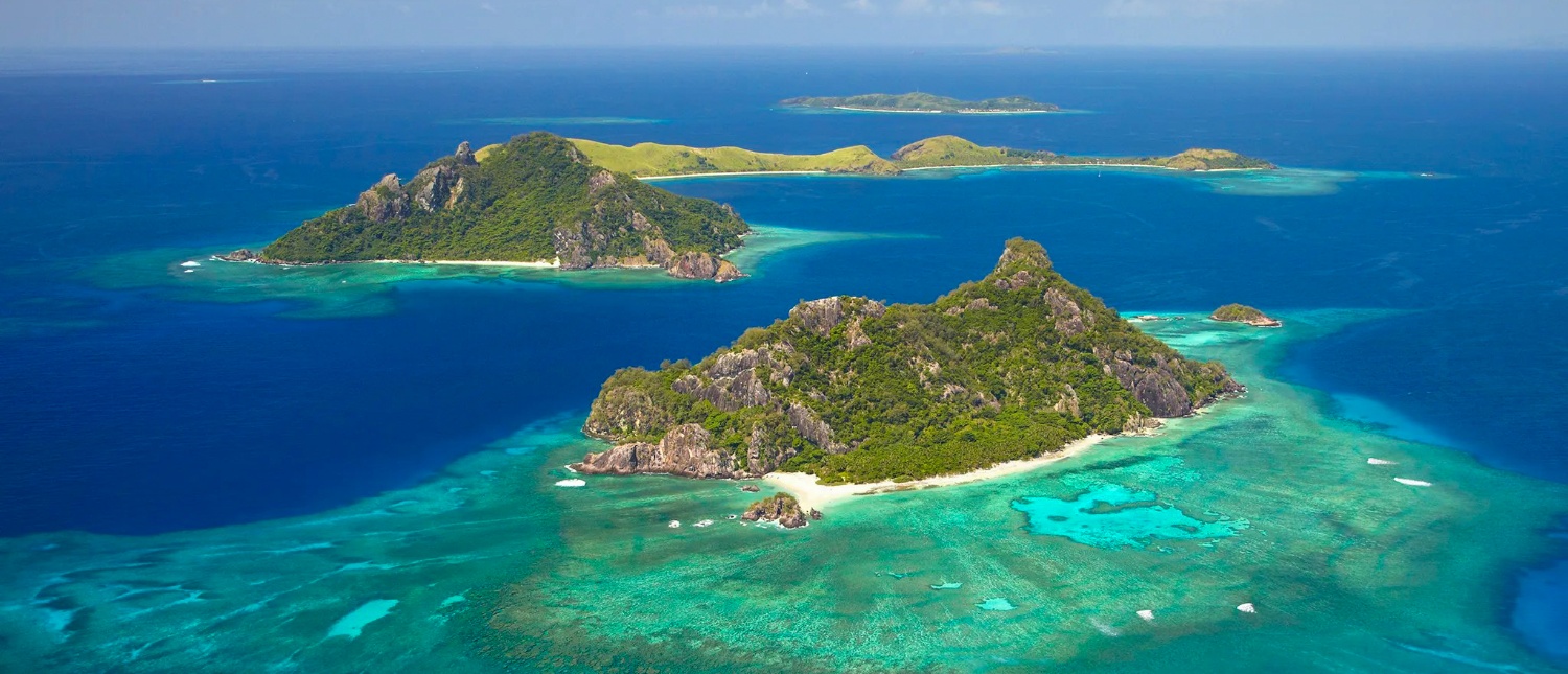 Lush green tropical islands emerge from clear turquoise waters, revealing vibrant coral reefs in Fiji.