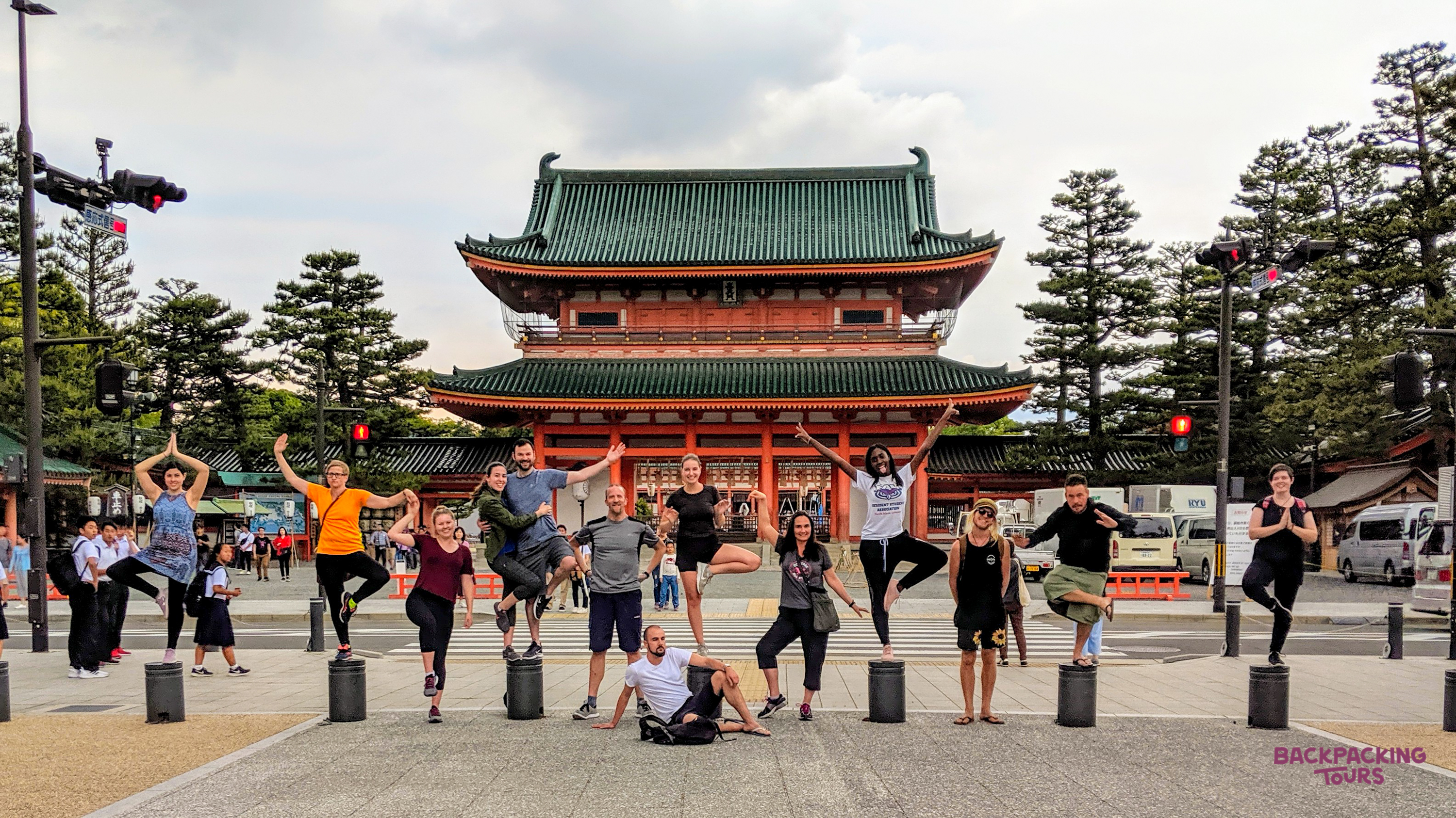 A group of joyful backpackers balancing on pillars in front of a grand traditional Japanese temple gate.