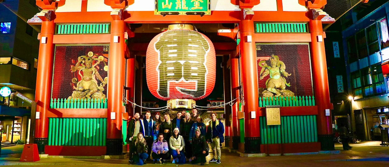 A group of happy travelers pose at night in front of Tokyo's illuminated Senso-ji Temple gate.