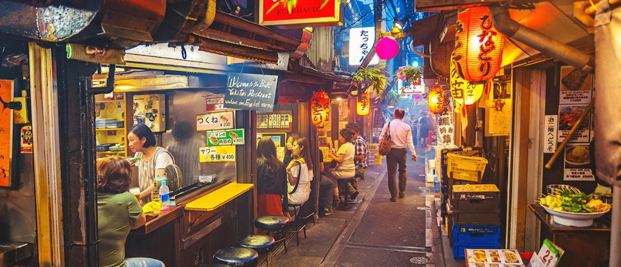 People enjoy food and drinks at small eateries in a lively, lantern-lit Tokyo alleyway at night.