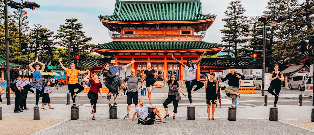 Joyful backpackers balance on bollards and jump playfully in front of a grand Japanese temple in Osaka.