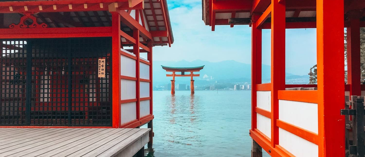 Red torii gate stands in calm water, framed by red wooden temple structures on Miyajima Island.
