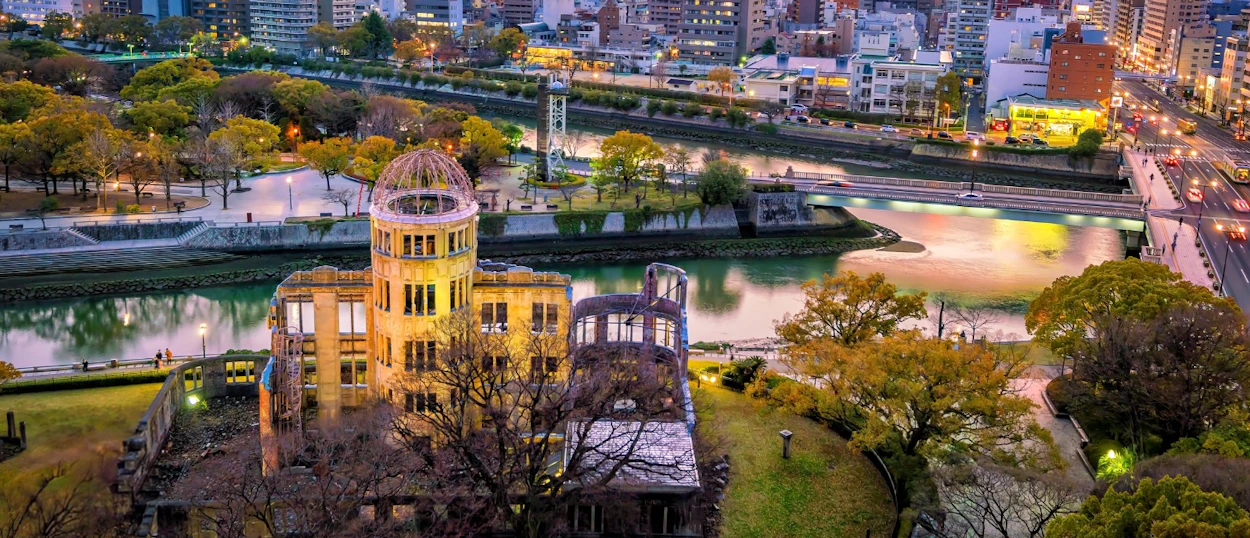The Hiroshima Peace Memorial stands by a river at dusk, surrounded by autumn trees and city lights.