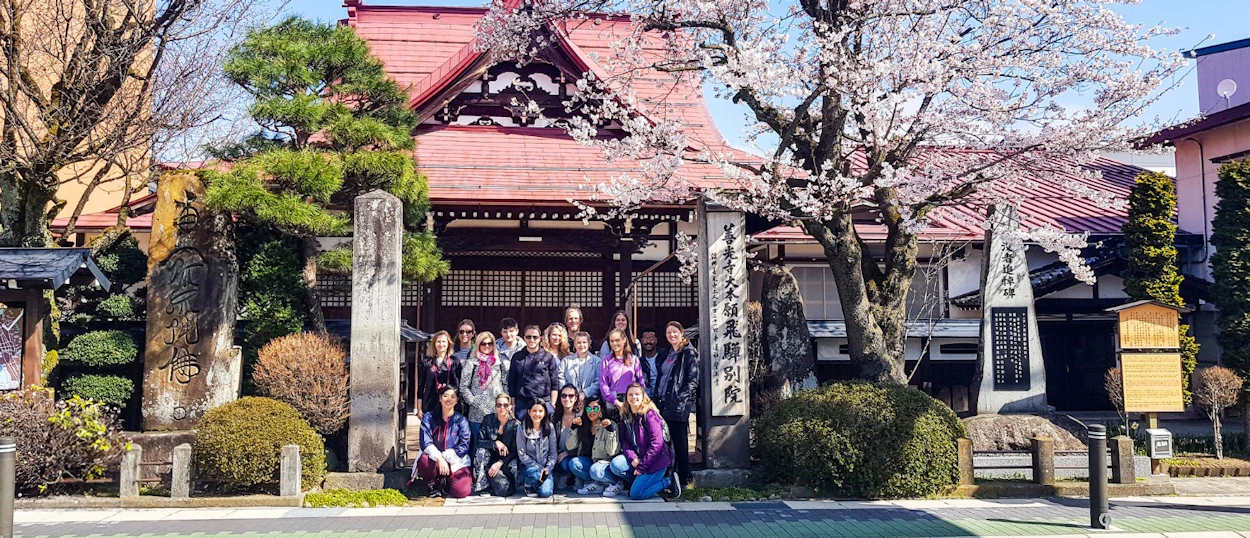 A happy group of travelers poses in front of a traditional Japanese temple in Hiroshima with cherry blossoms.