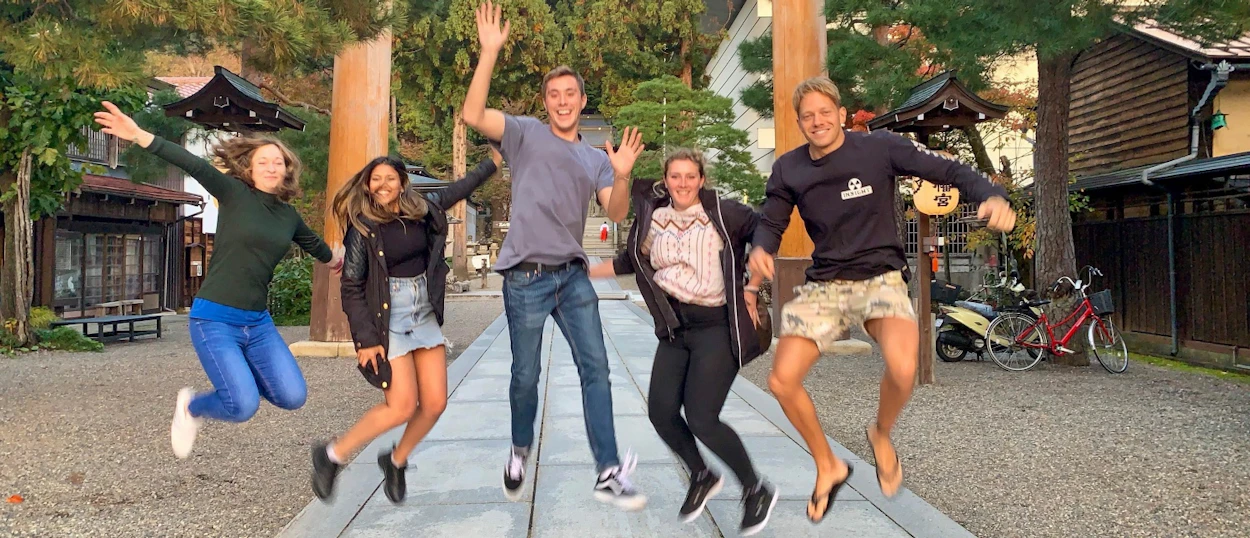 Five joyful friends jump high on a stone path at a traditional Japanese shrine in Tokyo.