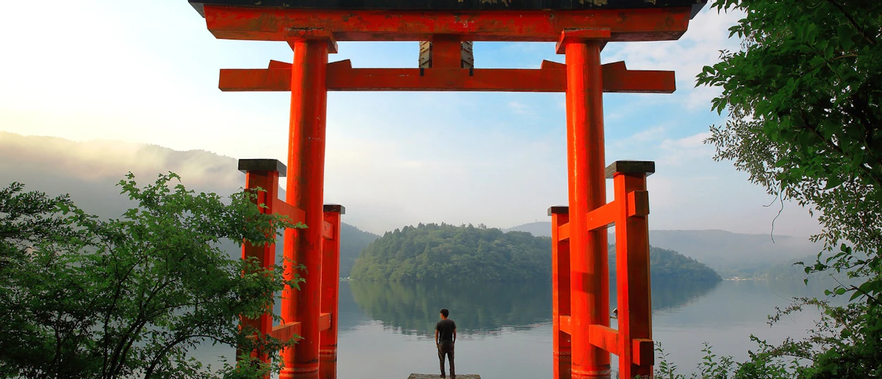A person stands at a red torii gate, looking over a calm lake and forested island in Hakone.