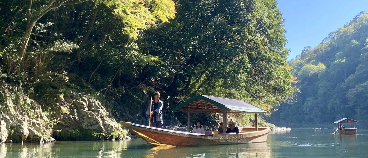 A traditional Japanese boat with passengers glides along a calm river surrounded by green mountains.