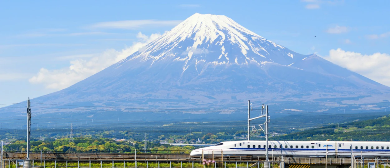 A white Shinkansen bullet train travels past the iconic, snow-capped Mount Fuji under a clear blue sky.