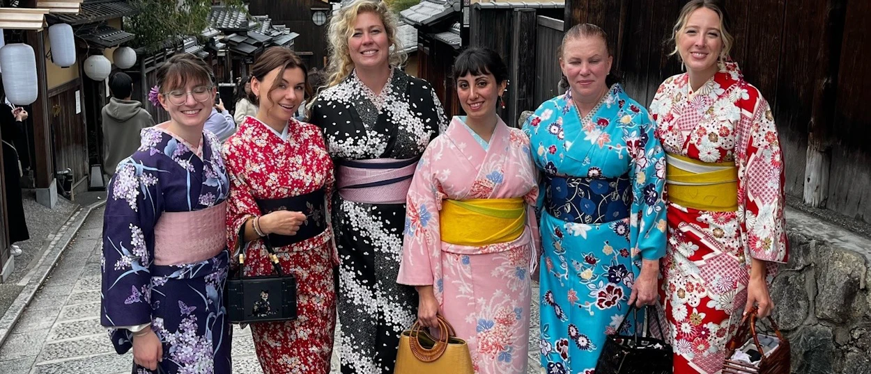 Six happy women in colorful kimonos pose on a traditional lantern-lit street in Kyoto.